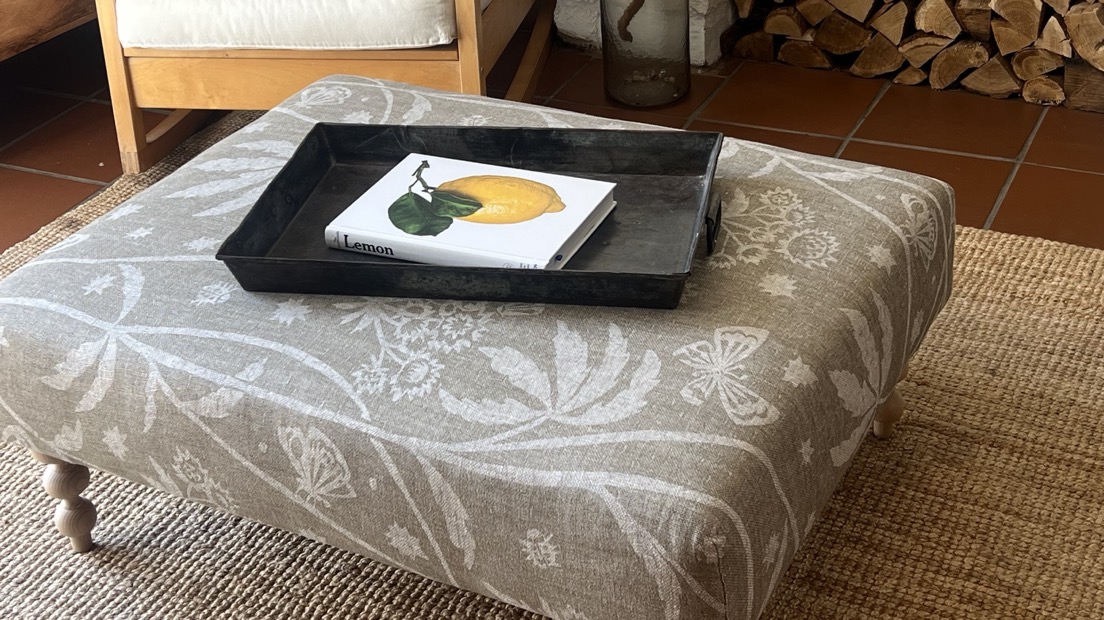 A linen-covered footstool holding a metal tray, in a room with a white brick fireplace and stacked logs